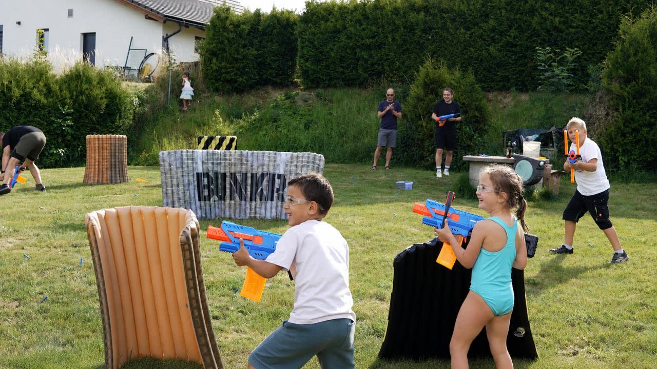 Děti i dospělí hrají pěnovou blaster bitvu mezi nafukovacími překážkami